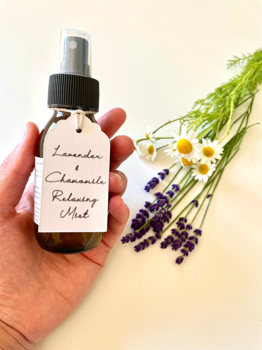 Lavender pillow spray handmade in Ireland. The brown glass bottle is held in a hand beside fresh lavender and chamomile flowers.