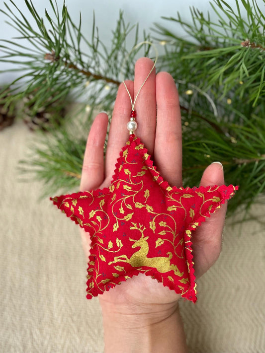 Red star fabric Christmas ornament with gold deer and holly pattern. Finished with glass beads and a faux pearl, it is shown on a hand