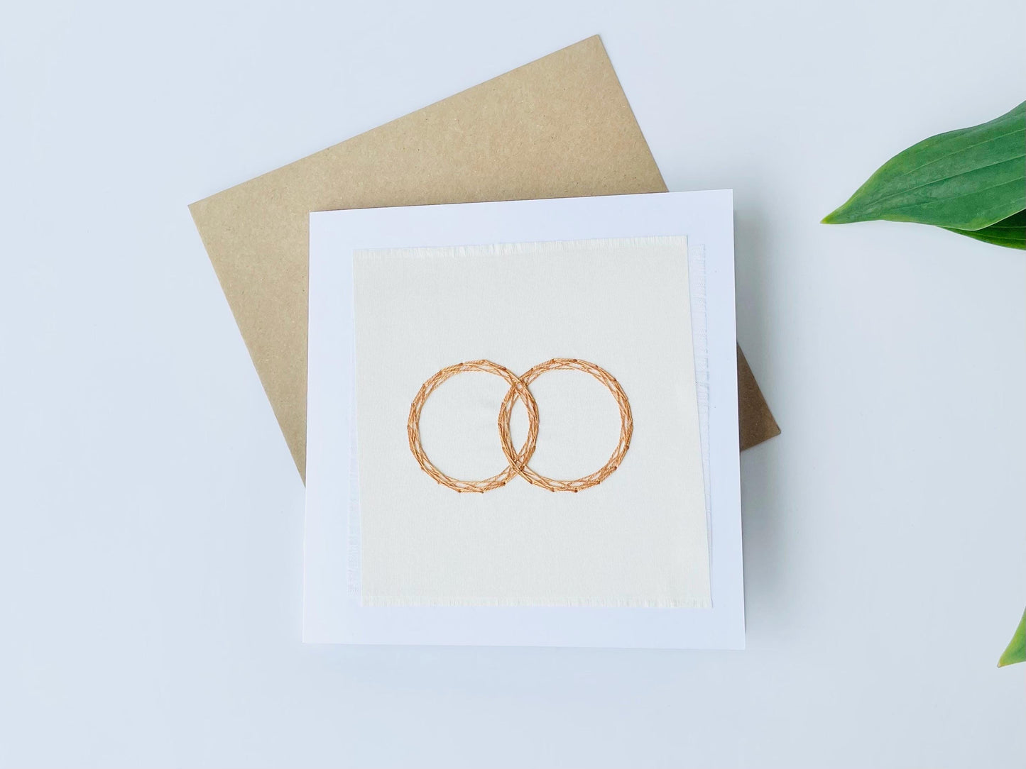 Hand embroidered wedding greeting card in white with two interlocking gold rings stitched onto fabric on the front.