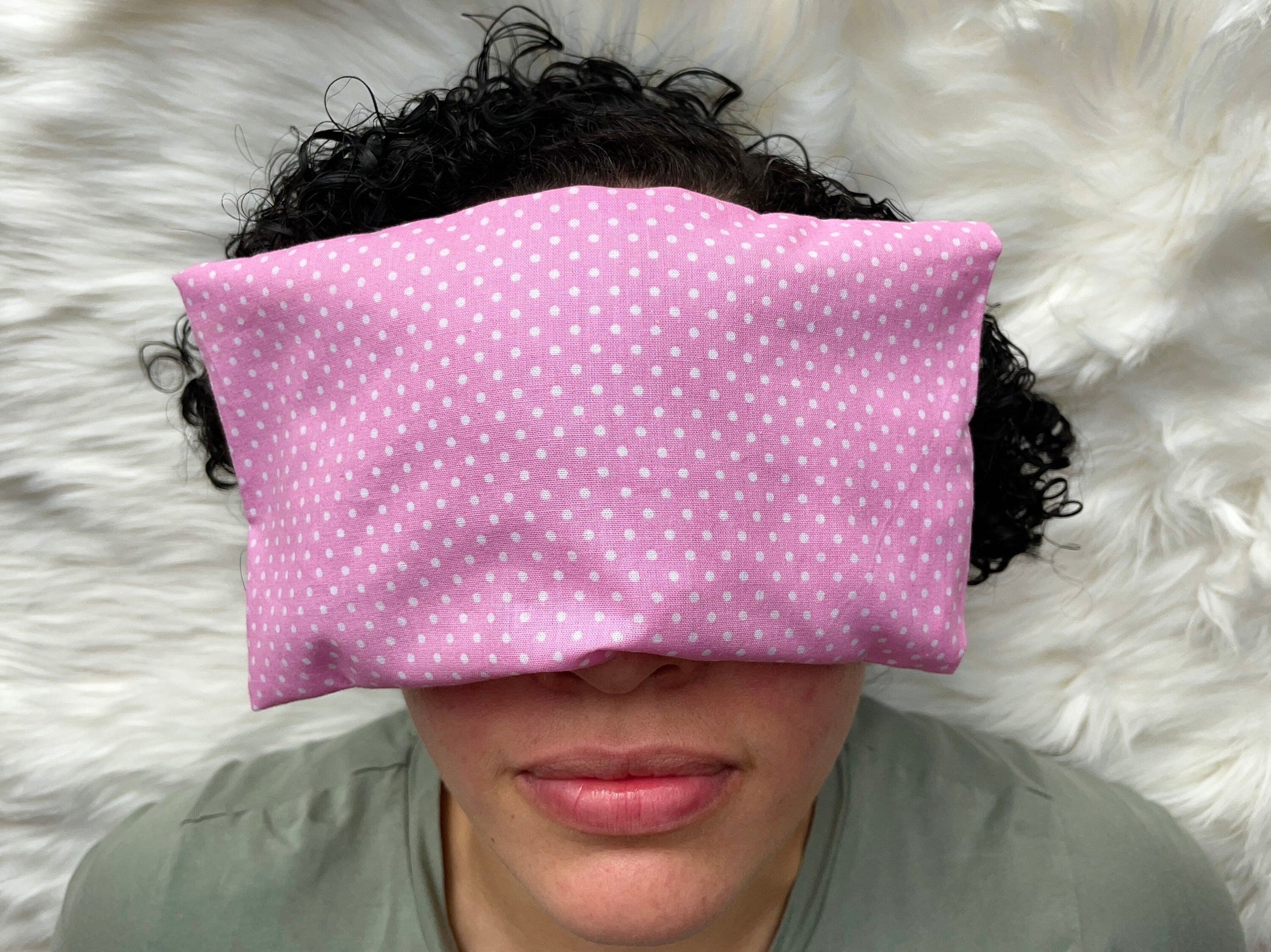 Pink weighted eye pillow with white polka dots shown on a female model with dark curly hair.