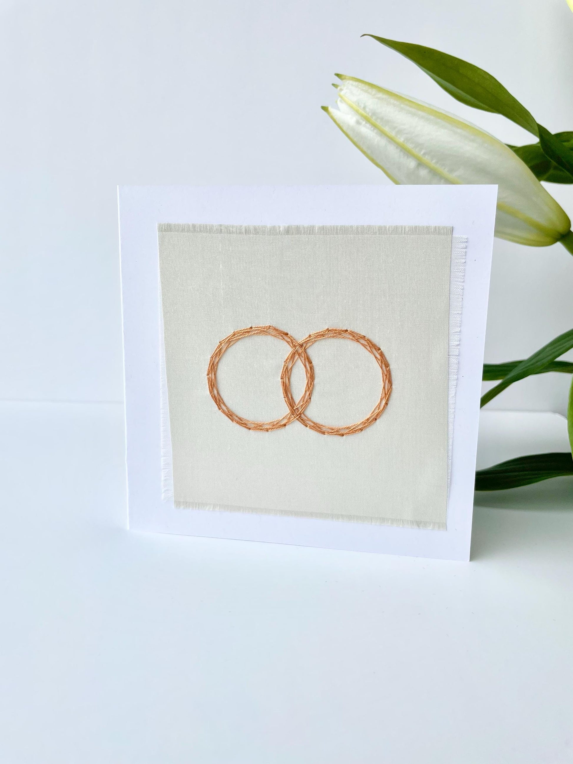 Textile wedding day greeting card hand stitched in Ireland, white card showing two interlocking gold rings embroidered in a geometric style 